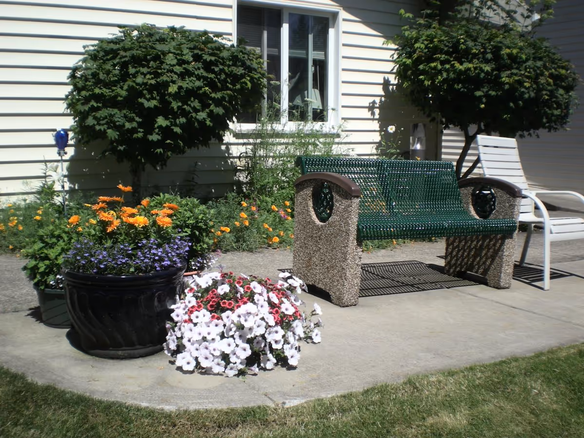 Outdoor patio area with a green metal bench and a white chair on a concrete surface. There are two small trees and several flower pots with colorful flowers in front of a light-colored building with a window.