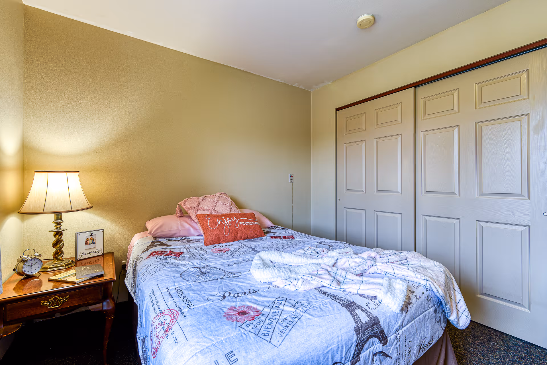 Small bedroom with a single bed covered in patterned bedding, a bedside table with a lamp and alarm clock, and sliding closet doors.