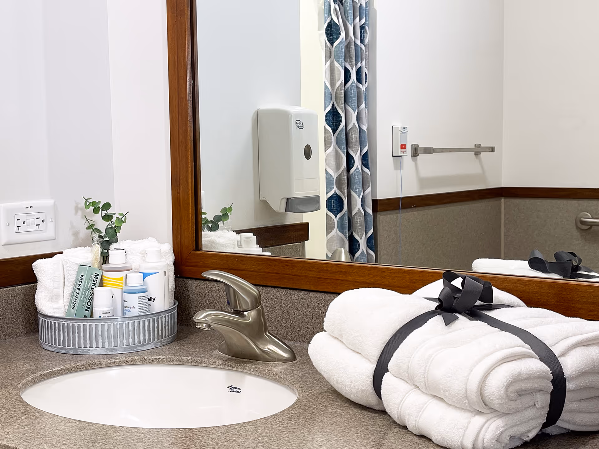 A clean bathroom sink area with a silver faucet, a large wooden-framed mirror, a metal tray holding rolled white towels and various toiletries, and a neatly folded stack of white towels tied with a black ribbon. In the mirror's reflection, a soap dispenser, a patterned shower curtain, a towel rack, and a grab bar are visible.