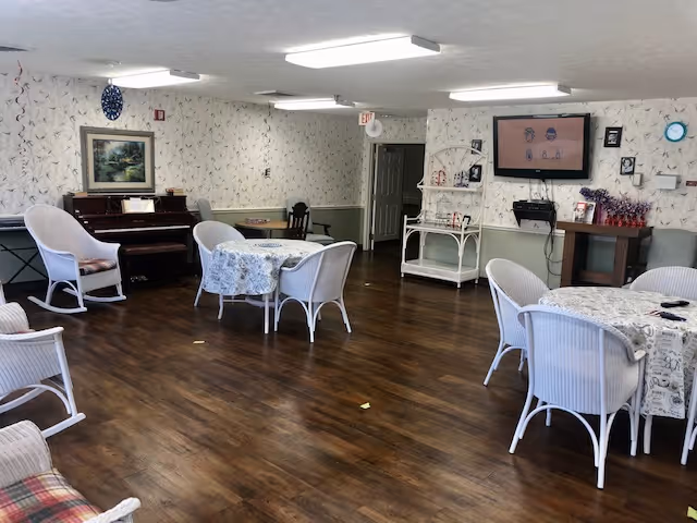 A cozy common area with wooden flooring, several round tables covered with patterned tablecloths, and white wicker chairs. There is a piano against the wall with a framed painting above it, a TV mounted on the wall, and a small white shelving unit. The walls have a light floral wallpaper, and the room is lit by ceiling lights.