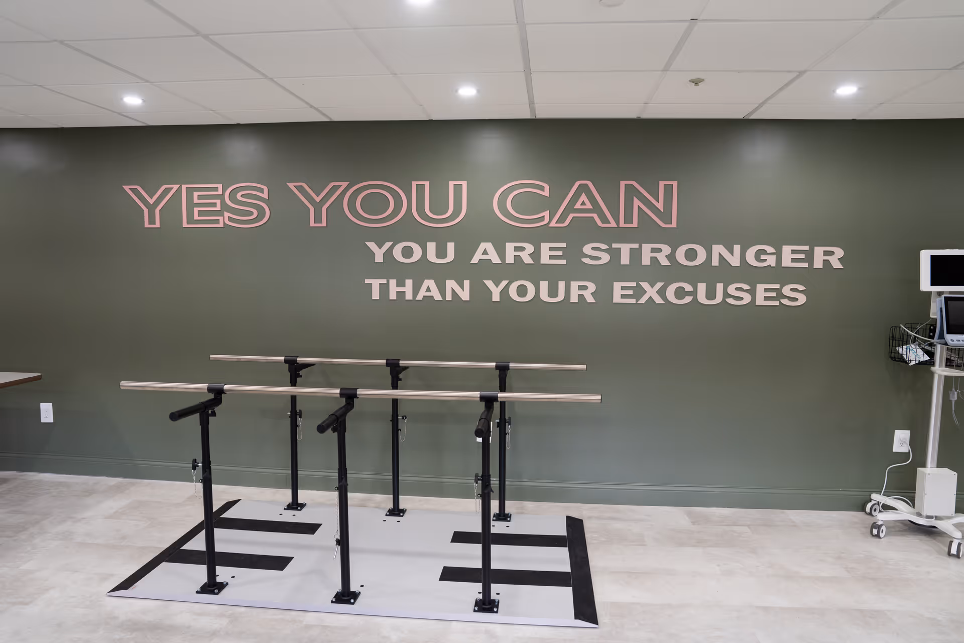 Rehabilitation exercise room with parallel bars in front of a green wall reading 'YES YOU CAN YOU ARE STRONGER THAN YOUR EXCUSES'.