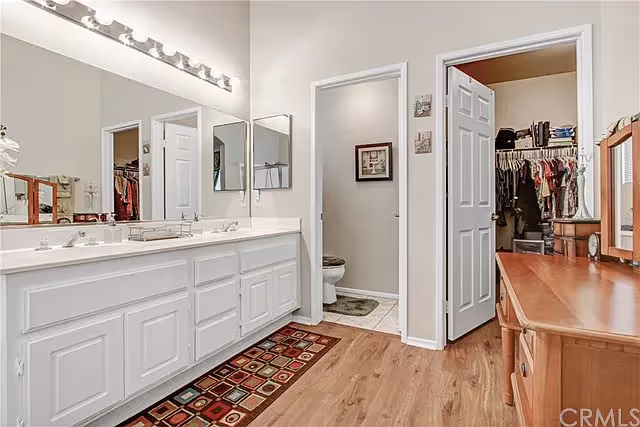Bathroom with a long double-sink vanity, large mirror, hardwood floor, and open doors to a toilet room and walk-in closet.
