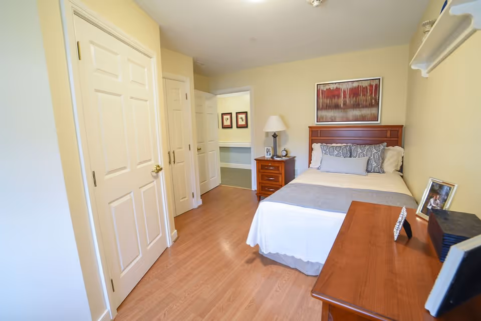 A cozy bedroom in a senior living facility featuring a single bed with white and gray bedding, a wooden headboard, and decorative pillows. Next to the bed is a wooden nightstand with a lamp, clock, and framed photo. Across from the bed is a wooden dresser with framed photos and decorative items. The room has light-colored walls, wooden flooring, and two closed white doors on the left side. A doorway leads to another room with framed artwork visible on the wall.