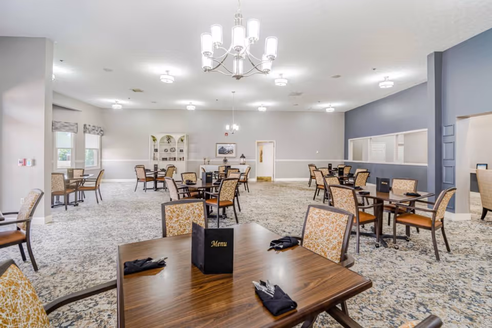 A spacious dining room with multiple wooden tables and chairs arranged neatly. Each table has a menu and black napkins with silverware. The room has light-colored walls, patterned carpet, several ceiling lights, and a chandelier. There are windows on the left side and a decorative shelf with plates and other items at the far end.