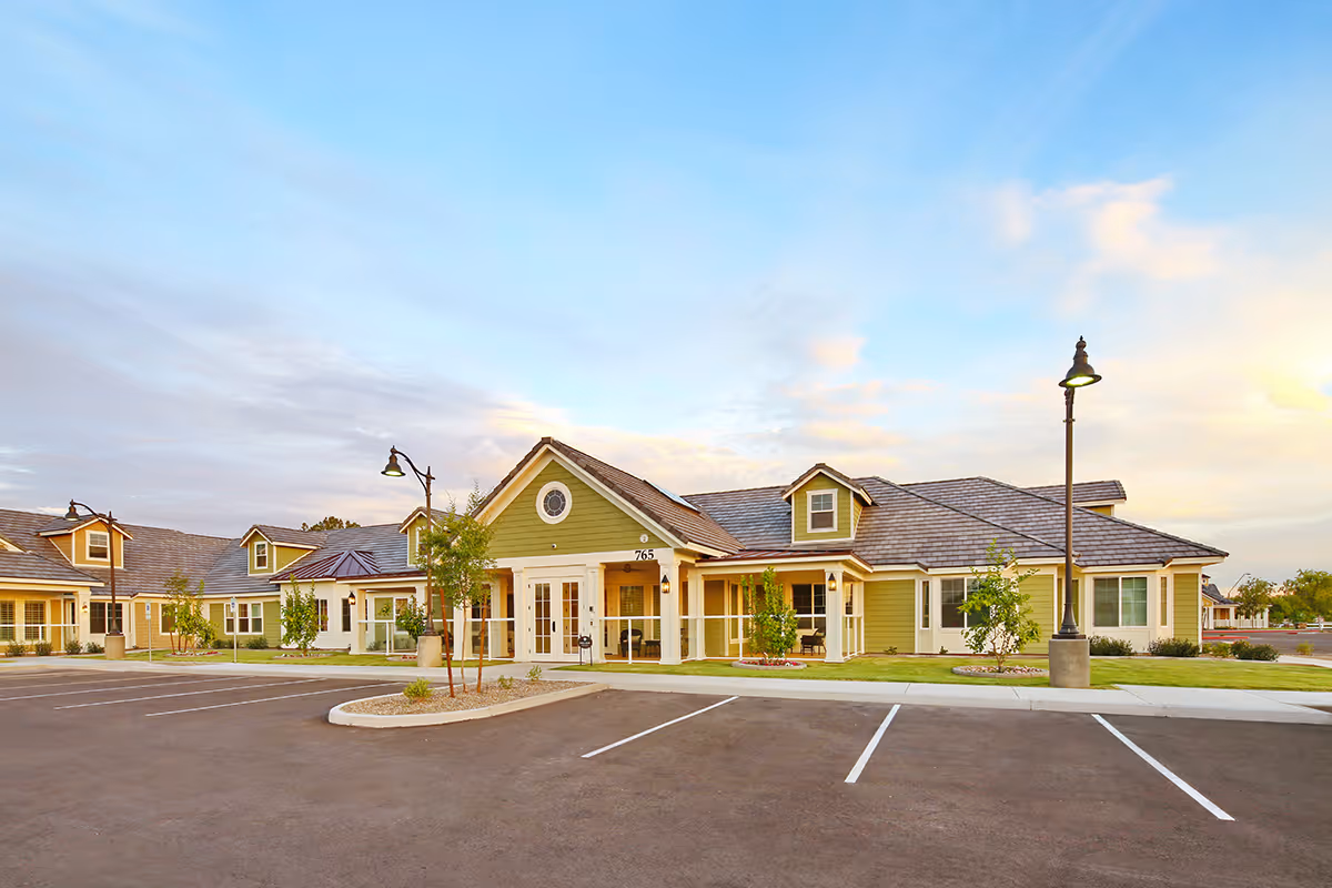 Single-story yellow-green assisted living building with a wide parking lot and lamp posts under a blue sky.