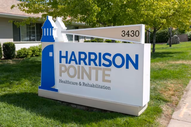 Outdoor sign for Harrison Pointe Healthcare & Rehabilitation with a blue lighthouse graphic and the address number 3430, situated on a grassy area with trees and a building in the background.