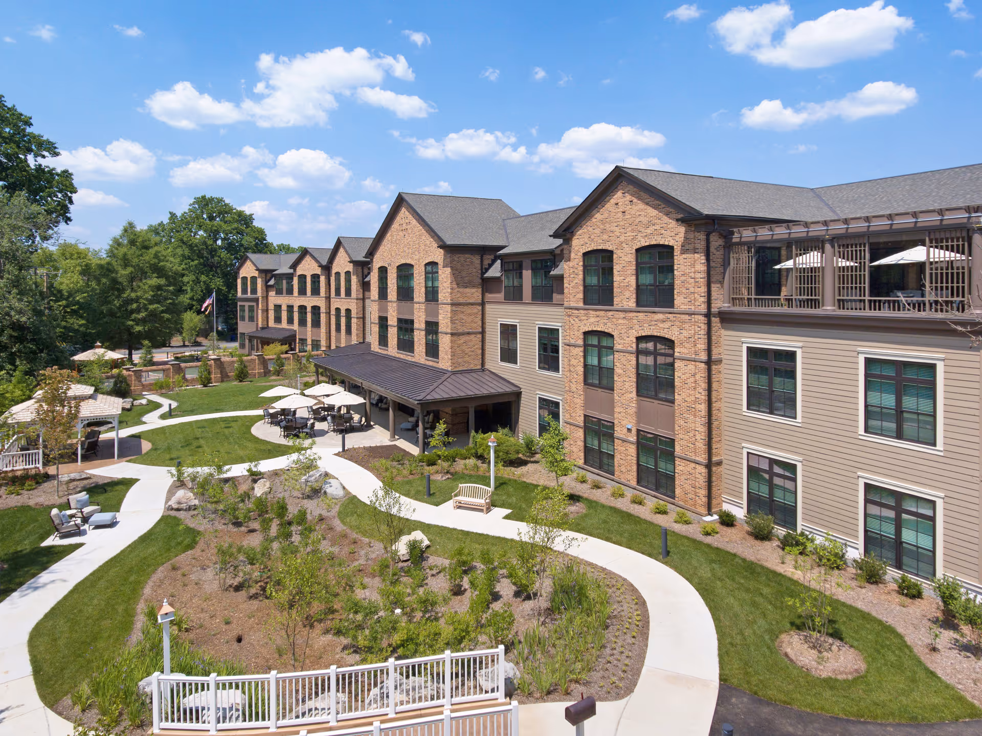 Exterior view of a senior living facility named Sunrise of McLean Village, showing a large brick and siding building with multiple windows, surrounded by landscaped gardens, walking paths, outdoor seating areas with tables and umbrellas, and green lawns under a blue sky with scattered clouds.