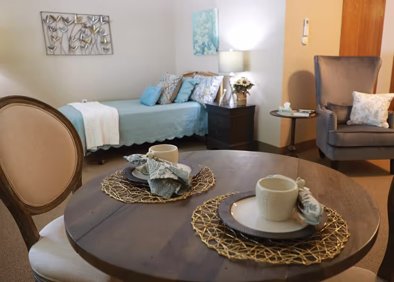 A cozy senior living suite showing a round dining table set for two in the foreground and a bed, nightstand with lamp, and armchair in the background.