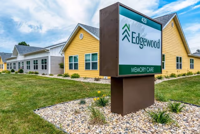 Exterior view of Edgewood Memory Care Watertown facility showing a large sign with the facility name and address number 420. The building has yellow and gray siding with multiple windows, surrounded by a landscaped area with grass, rocks, and small plants under a partly cloudy sky.