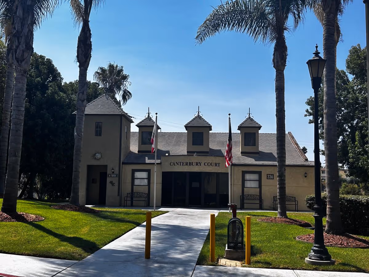Front entrance of a beige building labeled "Canterbury Court" with American flags, palm trees, and a paved walkway leading to the door.