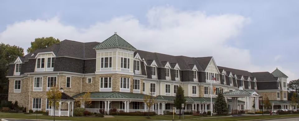 Exterior view of a large, multi-story senior living facility building with beige stone and white siding, green metal roofs on turrets, multiple windows, and a covered entrance. The sky is partly cloudy and there are trees and shrubs around the building.