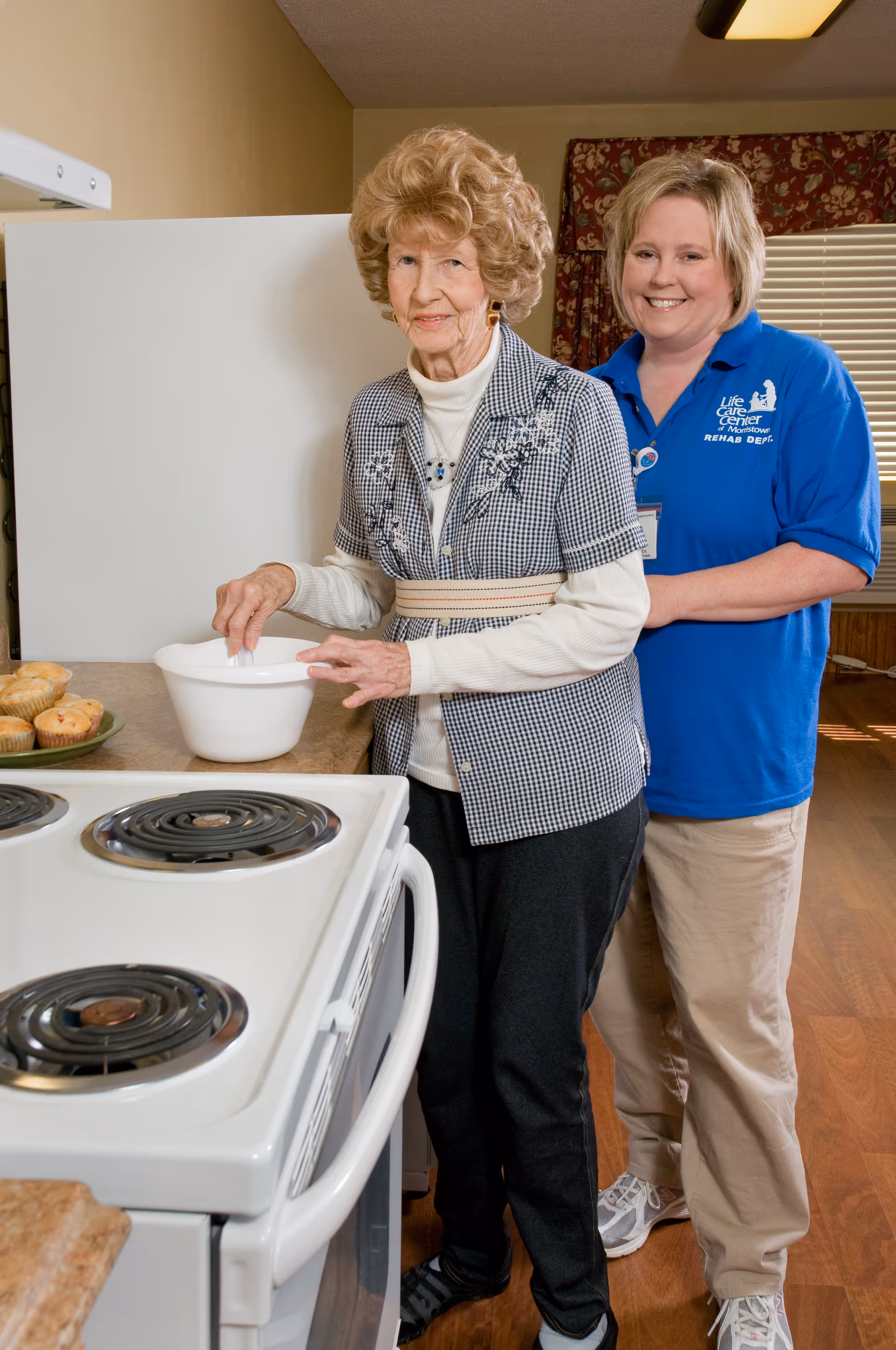 An elderly woman stirs a bowl on a kitchen counter while a smiling caregiver stands behind her.