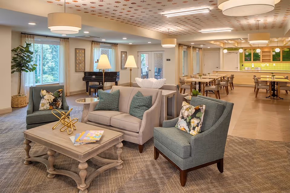 A bright and spacious senior living common area featuring comfortable seating with two armchairs and a loveseat around a wooden coffee table with books and decorative items. In the background, there is a dining area with tables and chairs, a green tiled backsplash kitchen area, and a black grand piano near windows with sheer curtains. The ceiling has patterned decorations and modern light fixtures.