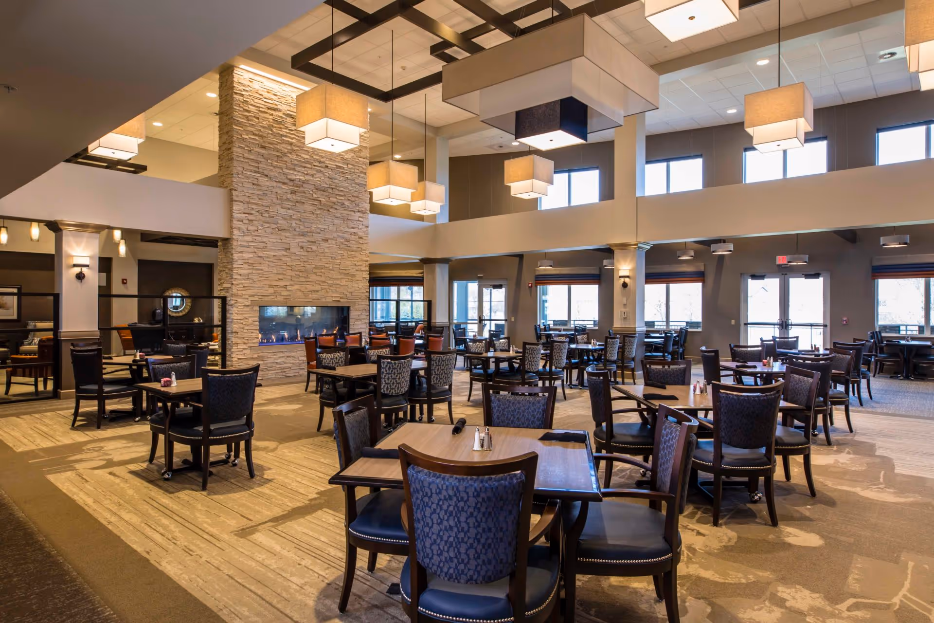 A spacious dining room with multiple wooden tables and chairs arranged neatly. The room features a tall stone fireplace in the center, modern hanging light fixtures, large windows allowing natural light, and a high ceiling with a grid pattern.