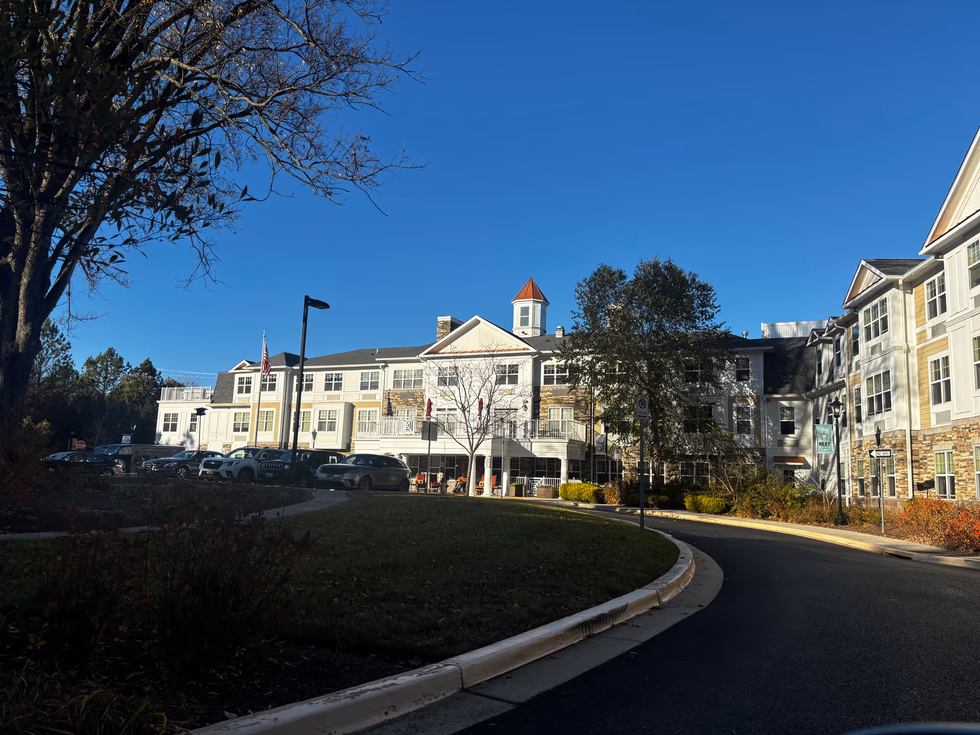 Front exterior of a multi-story senior living building with a curved driveway, parked cars, and trees under a clear blue sky.