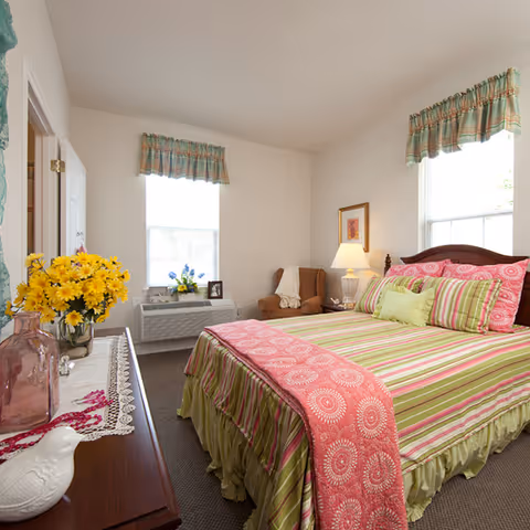 A bright and cozy bedroom with a bed dressed in green and pink striped bedding and a pink patterned throw. The room has two windows with plaid valances, a wooden dresser with decorative items including a vase of yellow flowers, and a comfortable armchair next to a side table with a lamp and framed artwork on the wall.