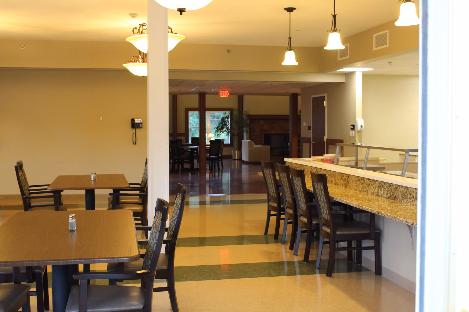 Interior view of a dining area in Jill's House Memory Care facility featuring wooden tables with chairs, a counter with high chairs, pendant lights hanging from the ceiling, and a seating area with armchairs and a fireplace in the background.