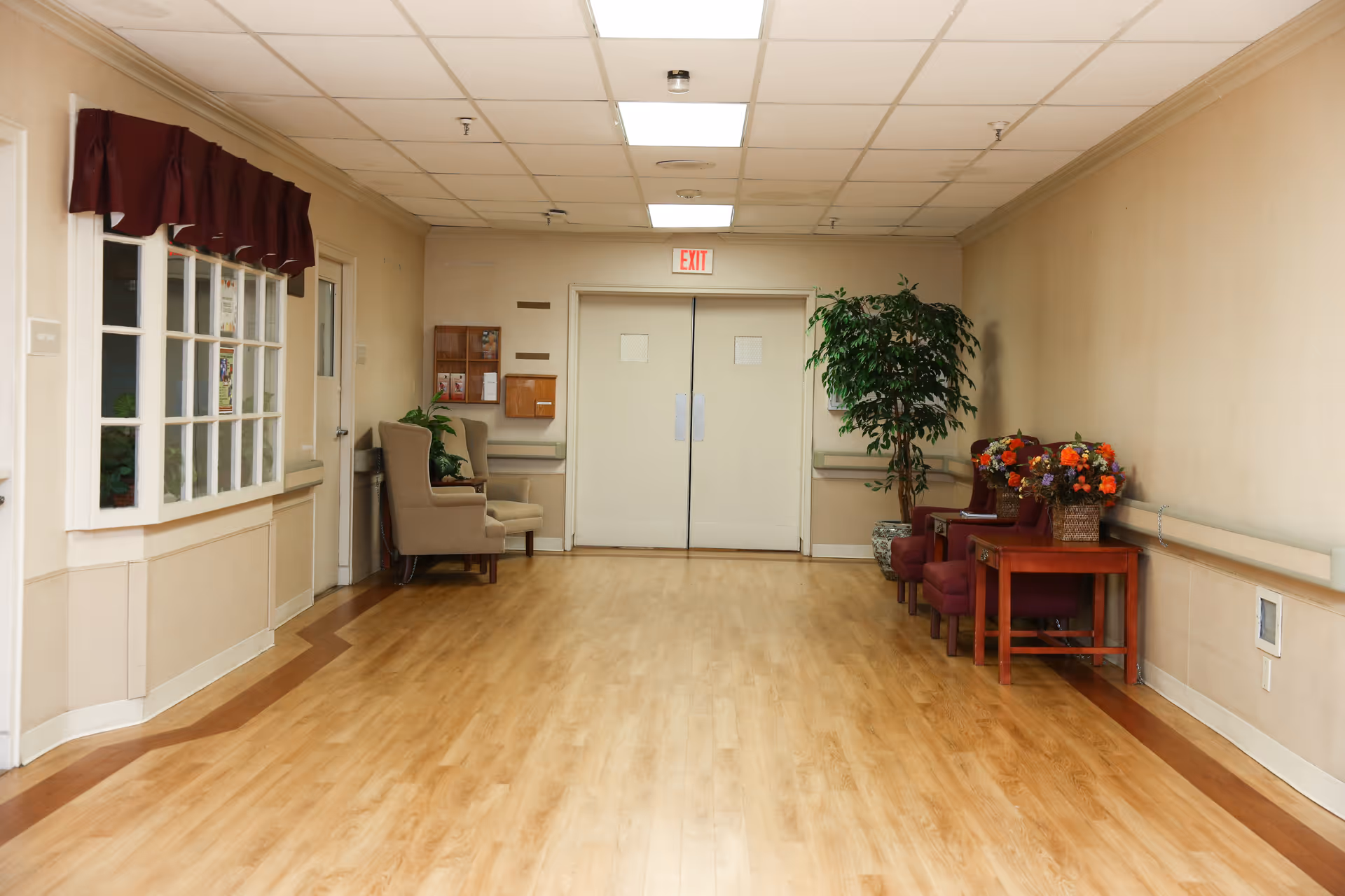 A clean and well-lit hallway in a senior living facility with light wood flooring, beige walls, and a white ceiling with fluorescent lights. On the left side, there is a window with a maroon valance and two beige armchairs with a small table and plant. On the right side, there are two maroon chairs, a wooden table with two baskets of colorful flowers, and a large potted plant. Double white doors with an exit sign above are at the end of the hallway.