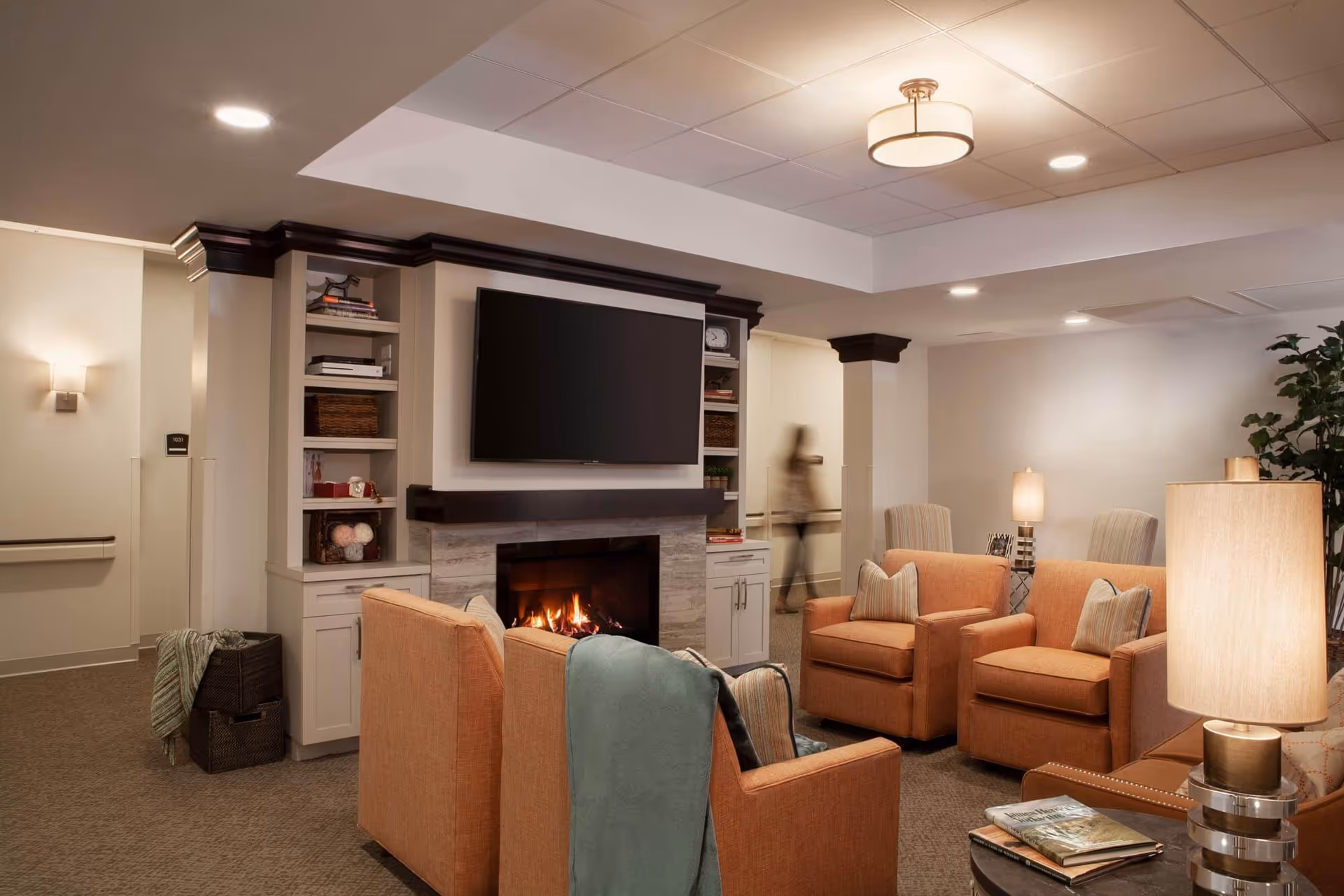 Cozy communal sitting area with a lit fireplace, wall-mounted TV, and several orange armchairs and lamps.