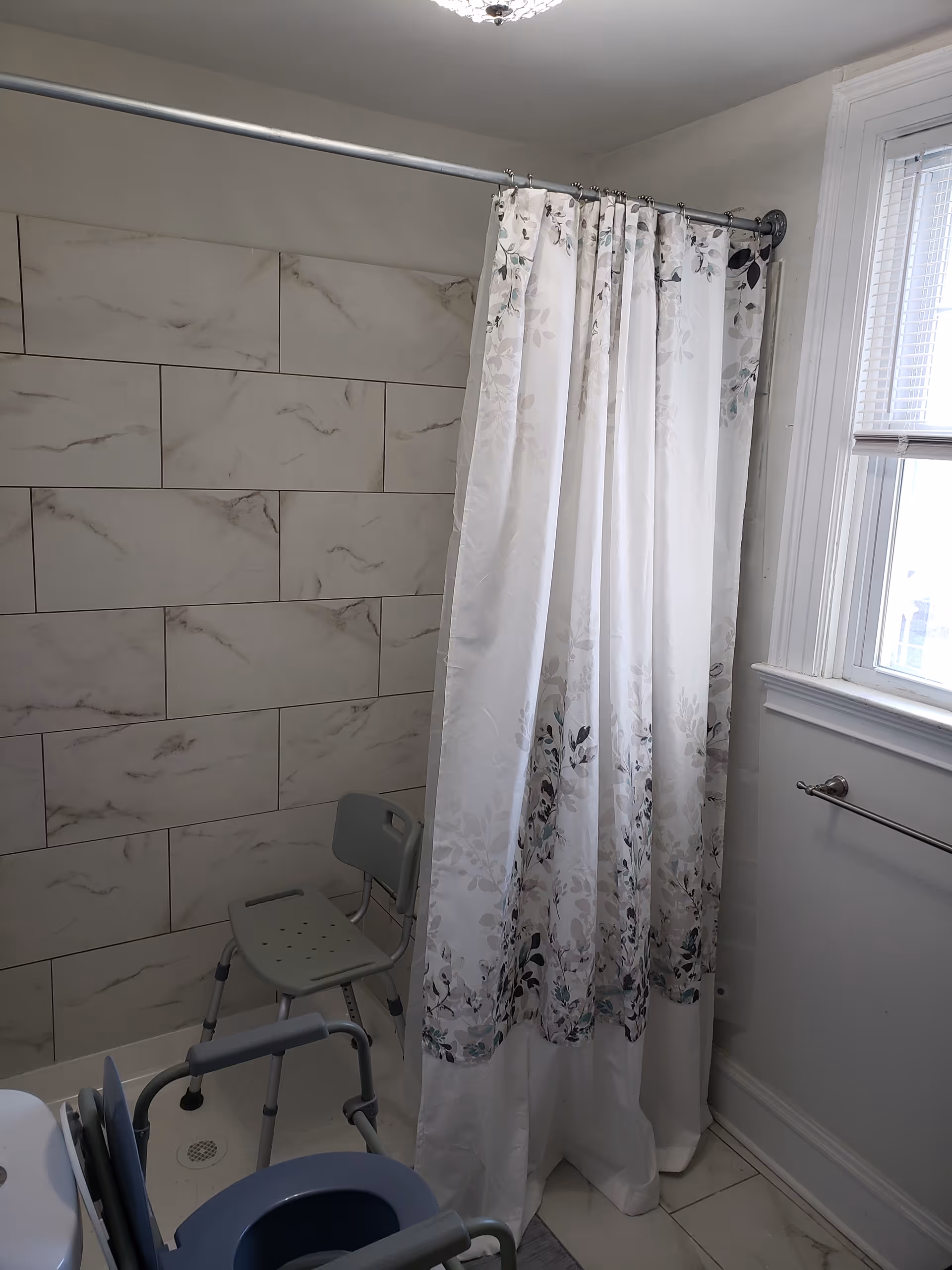 Bathroom with a tiled shower area featuring a white and gray floral shower curtain. Inside the shower is a gray shower chair. There is a window with blinds on the right side and a towel bar mounted on the wall below the window.