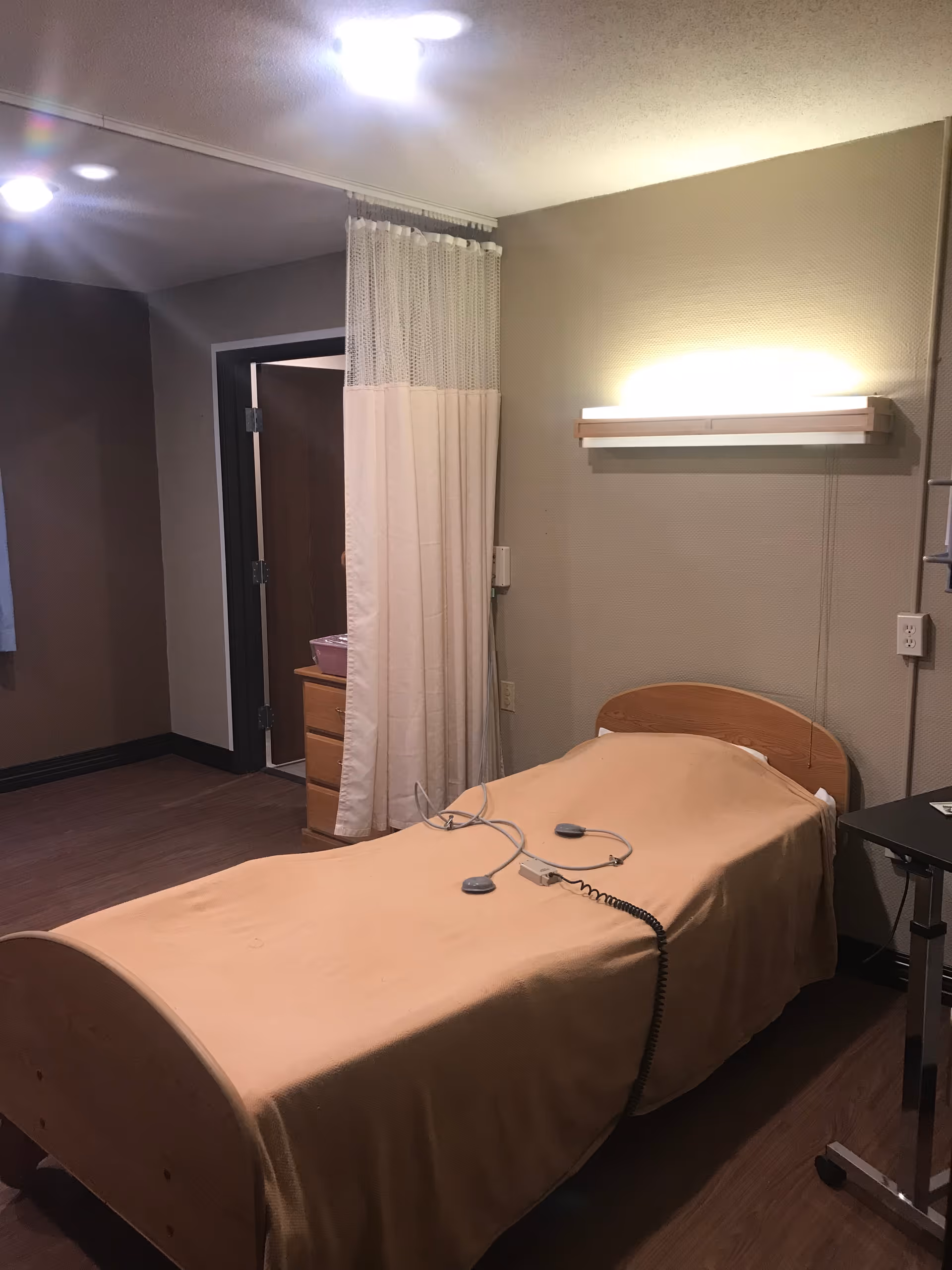 A single bed with a beige blanket in a senior living facility room. The bed has a call button device on it. There is a wall-mounted light above the bed and a privacy curtain partially drawn near the entrance to a bathroom. The room has wooden flooring and neutral-colored walls.
