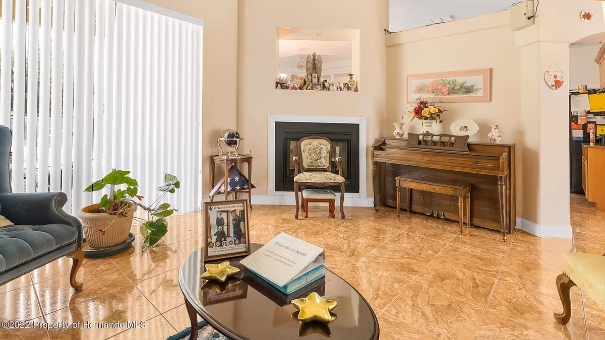 A cozy living room in an assisted living facility featuring a dark blue upholstered armchair, a potted plant, a wooden coffee table with a grievance book and decorative items, a vintage floral upholstered chair in front of a fireplace, and a wooden piano with floral decorations on top. The room has beige walls, tiled floors, vertical blinds covering a large window, and an open view into a kitchen area.