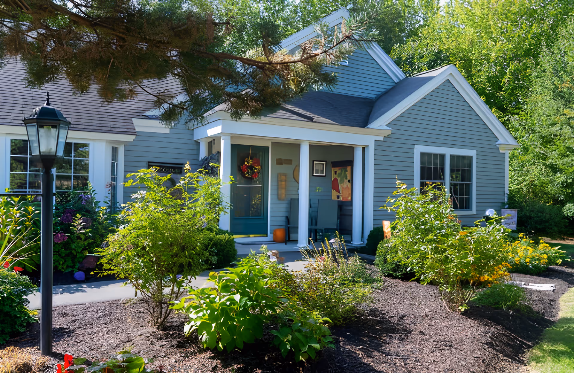 Front exterior view of a single-story house with blue siding and white trim, featuring a small covered porch with two white columns. The porch has a green door decorated with a wreath and two chairs. The house is surrounded by a well-maintained garden with various green shrubs, plants, and flowers. A black lamp post is visible on the left side of the image.