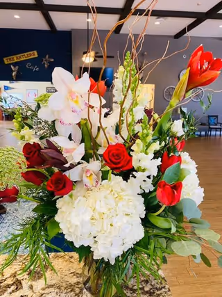A vibrant floral arrangement featuring white hydrangeas, red roses, white orchids, and red tulips placed on a granite countertop in a spacious room with wooden flooring and blue walls. In the background, there is a sign that reads 'THE ANTLERS Cafe' and some chairs and tables.