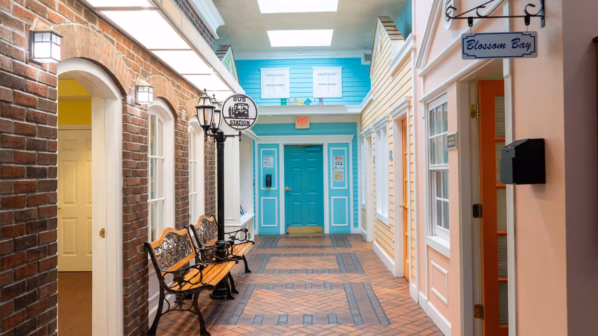 Indoor hallway designed to look like a small town street with colorful building facades, a brick wall, benches, a street lamp, and signs reading 'Bus Station' and 'Blossom Bay'. The floor is tiled in a brick pattern and there is a turquoise door at the end of the hallway.