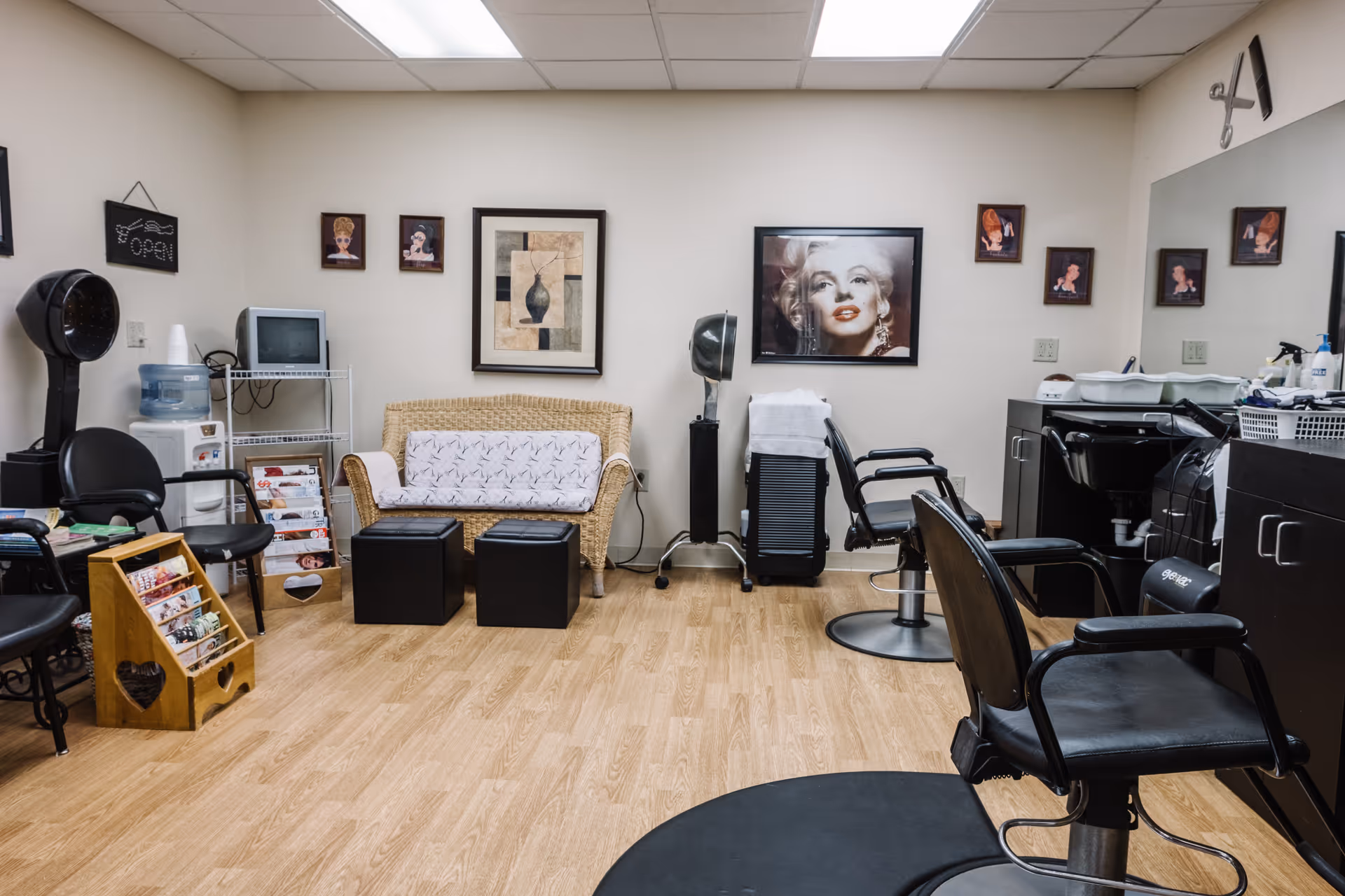 A small salon interior with styling chairs, a shampoo sink, drying hood, a wicker bench, magazine rack and framed wall art.