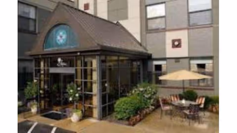 Entrance area of Vitality Living St. Matthews facility showing a glass-enclosed entryway with a peaked roof, surrounded by plants and shrubs. To the right, there is an outdoor seating area with a table, chairs, and a large umbrella. The building exterior is visible with multiple windows.