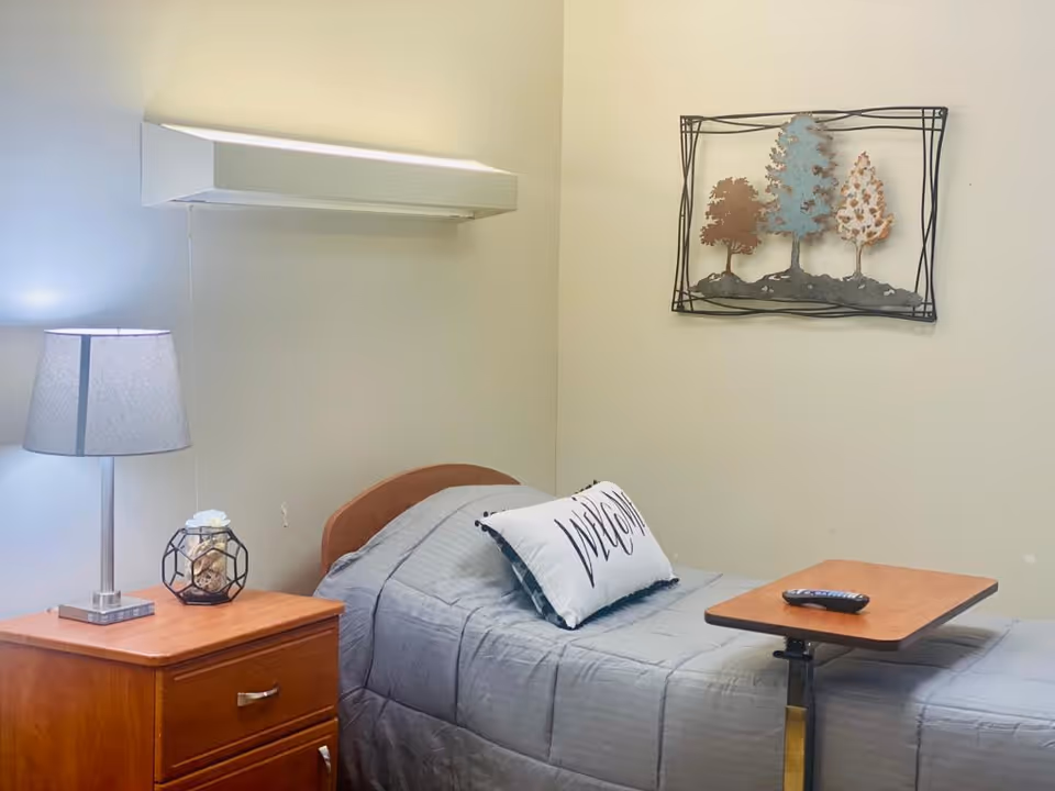 A small bedroom with a single bed covered in a gray quilt and a white pillow with the word 'Welcome' on it. Next to the bed is a wooden nightstand with a lamp and a decorative item. Above the bed on the wall is a metal wall art piece featuring three trees.