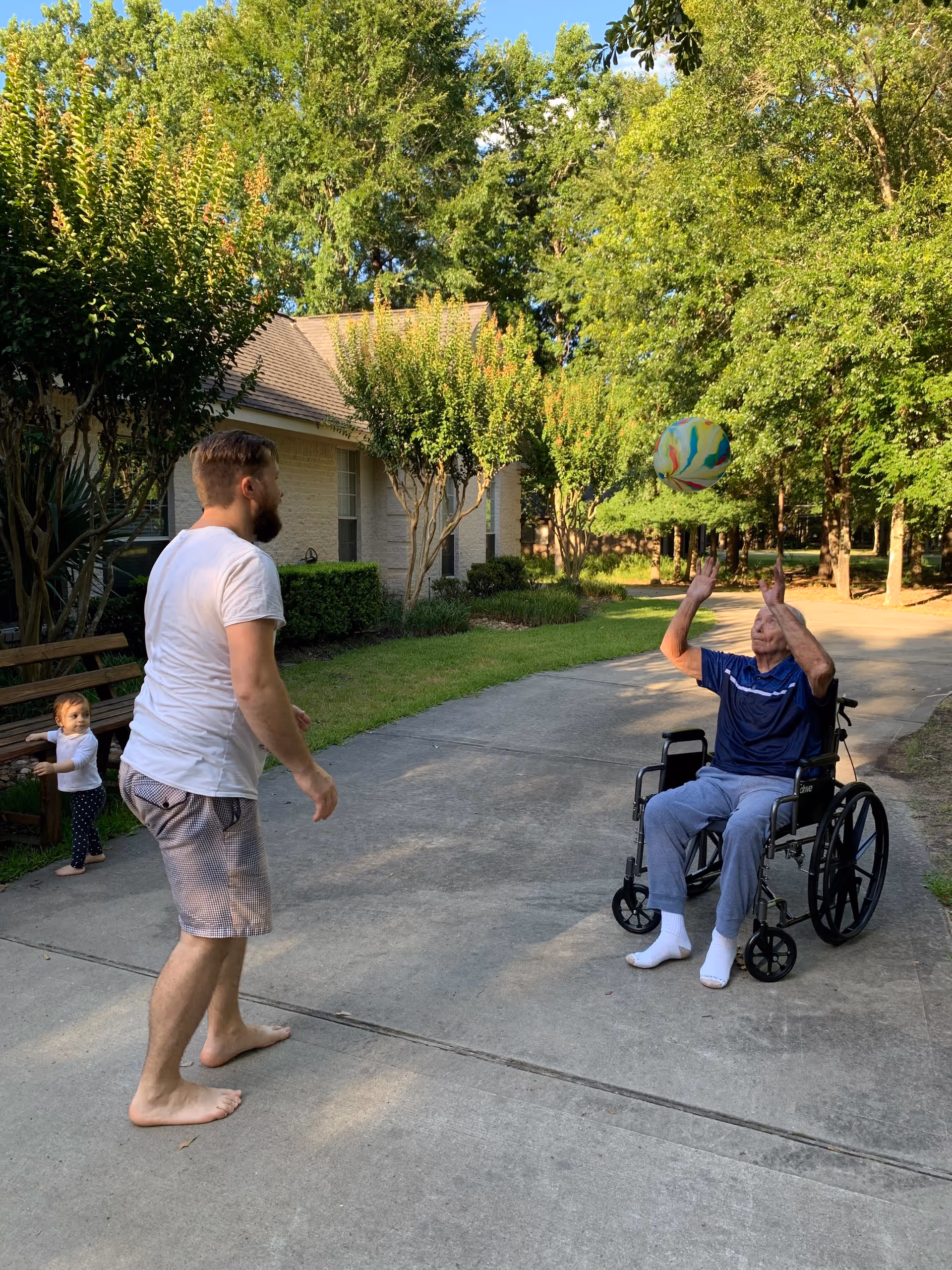 An elderly man in a wheelchair playing with a colorful ball outdoors on a paved pathway, accompanied by a barefoot man in shorts and a white t-shirt, with a small child standing near a bench in the background. The scene is surrounded by green trees and bushes near a residential building.