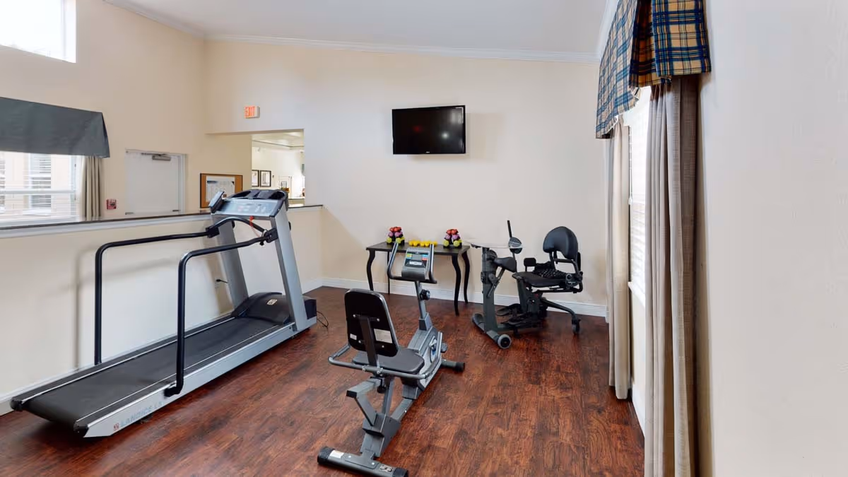 Small exercise room with a treadmill, recumbent bike, upright bike, a table of free weights and a wall-mounted TV.