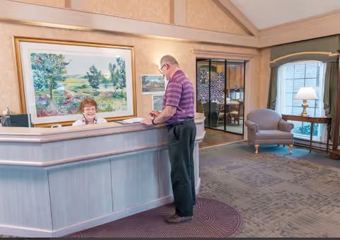 A man in a purple striped shirt stands at a reception desk speaking with a smiling receptionist seated behind the desk. The reception area features a large framed landscape painting on the wall, a patterned carpet, a comfortable armchair, a side table with a lamp, and a window with curtains.