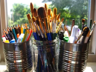 Paintbrushes and art supplies in metal cups sit on a sunlit windowsill.