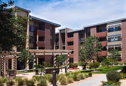 Outdoor courtyard area of a senior living facility with a pergola, benches, trees, shrubs, and a multi-story building in the background under a clear blue sky.
