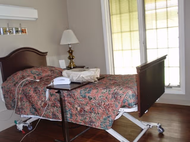 A hospital-style bed with a floral patterned bedspread in a room with wooden flooring. The bed has wheels and an adjustable frame. Next to the bed is a small overbed table with a white telephone and a beige bag on it. There is a brass lamp on a nightstand beside the bed and a large window with blinds letting in natural light.