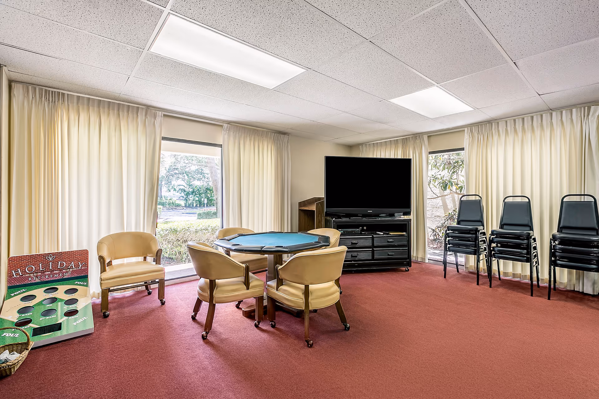 A common room with a red carpet, beige walls, and large windows covered with white curtains. The room contains a round table with four beige chairs on casters, a large flat-screen TV on a black stand, several stacks of black chairs, and a bean bag toss game board leaning against the wall.
