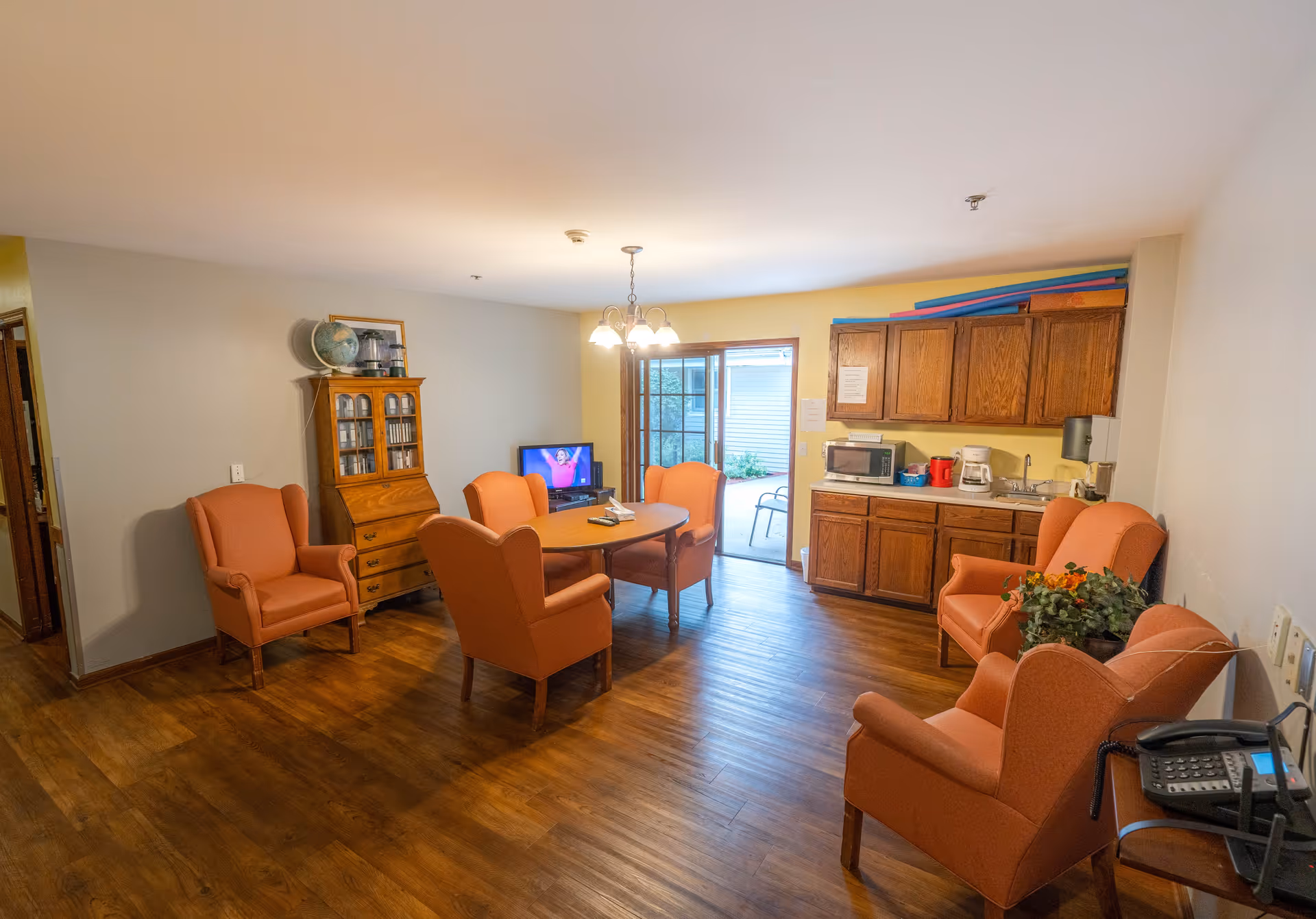 A cozy dining room at Park Hills West featuring a round table surrounded by four orange armchairs, a wooden cabinet, and a kitchenette with a microwave and sink. A door leads to an outdoor area.