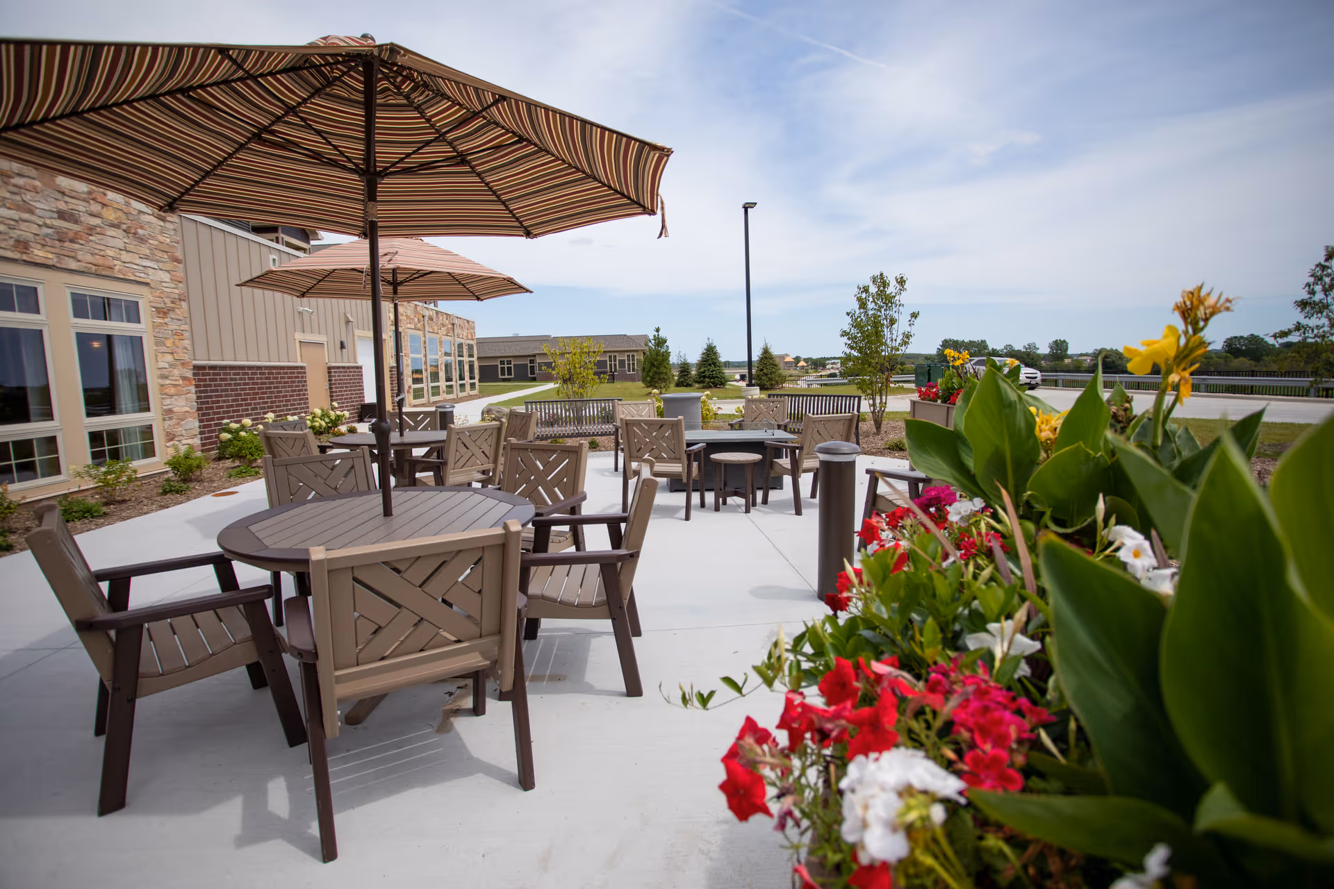 Outdoor patio area at a senior living facility with multiple round tables and chairs under large striped umbrellas. The patio is adjacent to a building with stone and brick exterior walls. There are colorful flowers and green plants in the foreground, and a clear sky with some clouds in the background.