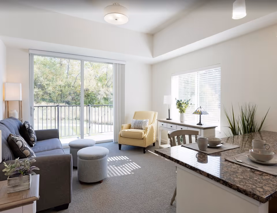 A bright and cozy living room in a senior living facility featuring a gray sofa with patterned pillows, two round gray ottomans, a yellow armchair with a decorative pillow, a white sideboard with a lamp and flowers, and a granite countertop with place settings. Large sliding glass doors and a window allow natural light to fill the room, with a view of greenery outside.