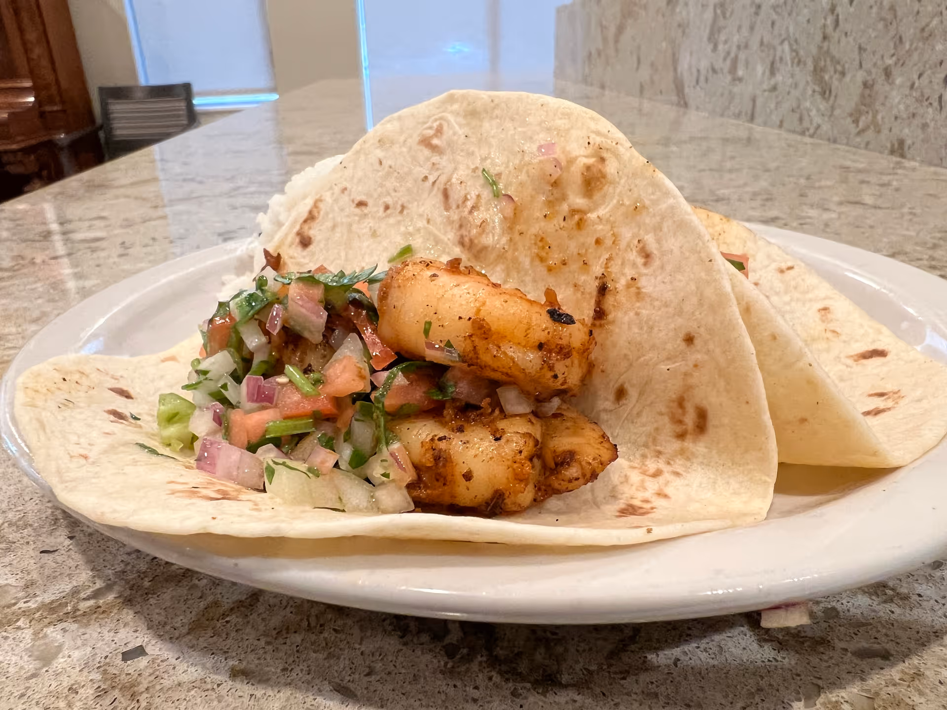 Two flour-tortilla tacos filled with grilled shrimp and diced pico de gallo on a white plate on a granite countertop.