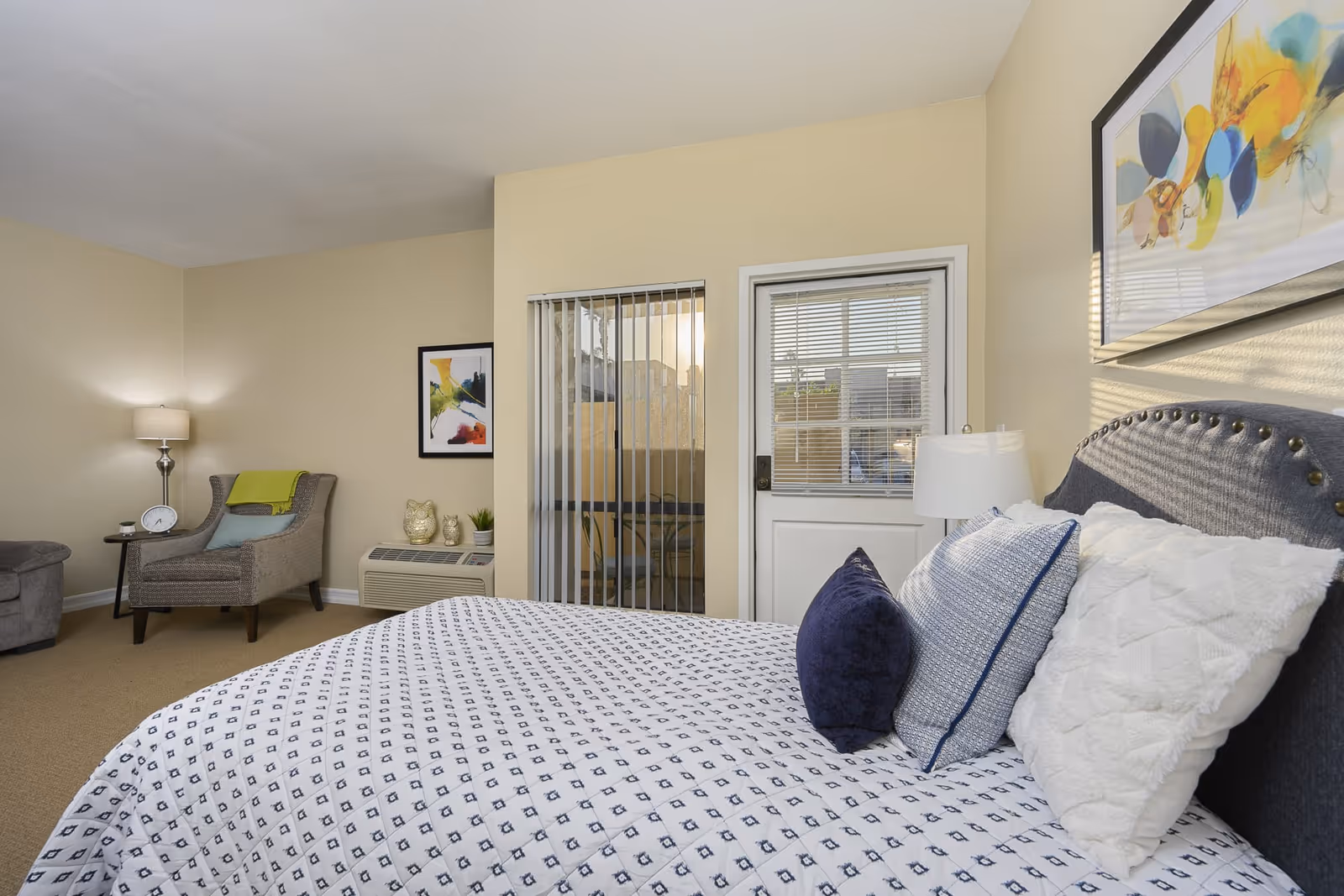 A cozy bedroom with a neatly made bed featuring white and blue patterned bedding and three pillows. To the right of the bed is a nightstand with a white lamp. On the wall above the bed is a colorful abstract painting. In the corner of the room, there is a gray armchair with a green throw blanket and a small side table with a clock and a small plant. Next to the armchair is an air conditioning unit with decorative owl figurines and a small plant on top. The room has beige walls and carpet, a sliding glass door with vertical blinds, and a white door with window blinds leading outside.