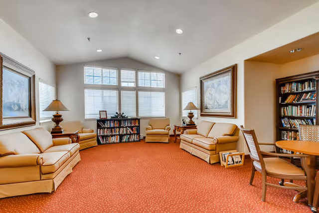 A spacious living room with beige sofas and armchairs arranged around the perimeter. The room features a red carpet, two wooden side tables with lamps, a bookshelf filled with books, and large windows with white blinds letting in natural light. There are framed paintings on the walls and a round wooden table with chairs on the right side.