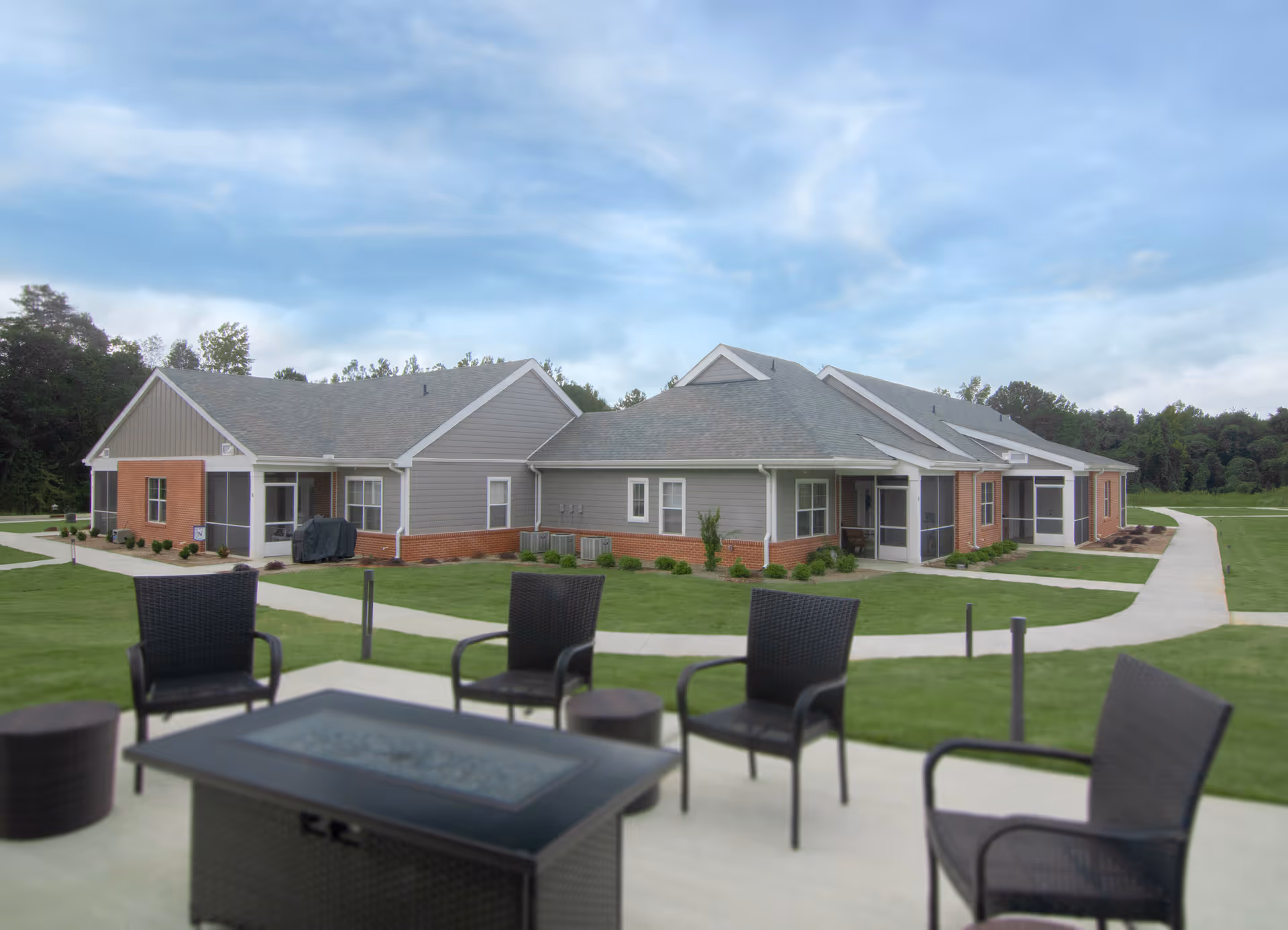 Single-story brick-and-siding retirement community building with walkways and outdoor patio chairs and a table in the foreground.