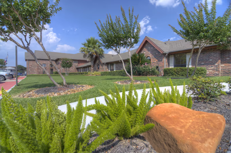 Front exterior of a brick senior living building with landscaped lawn, trees, and a walkway.