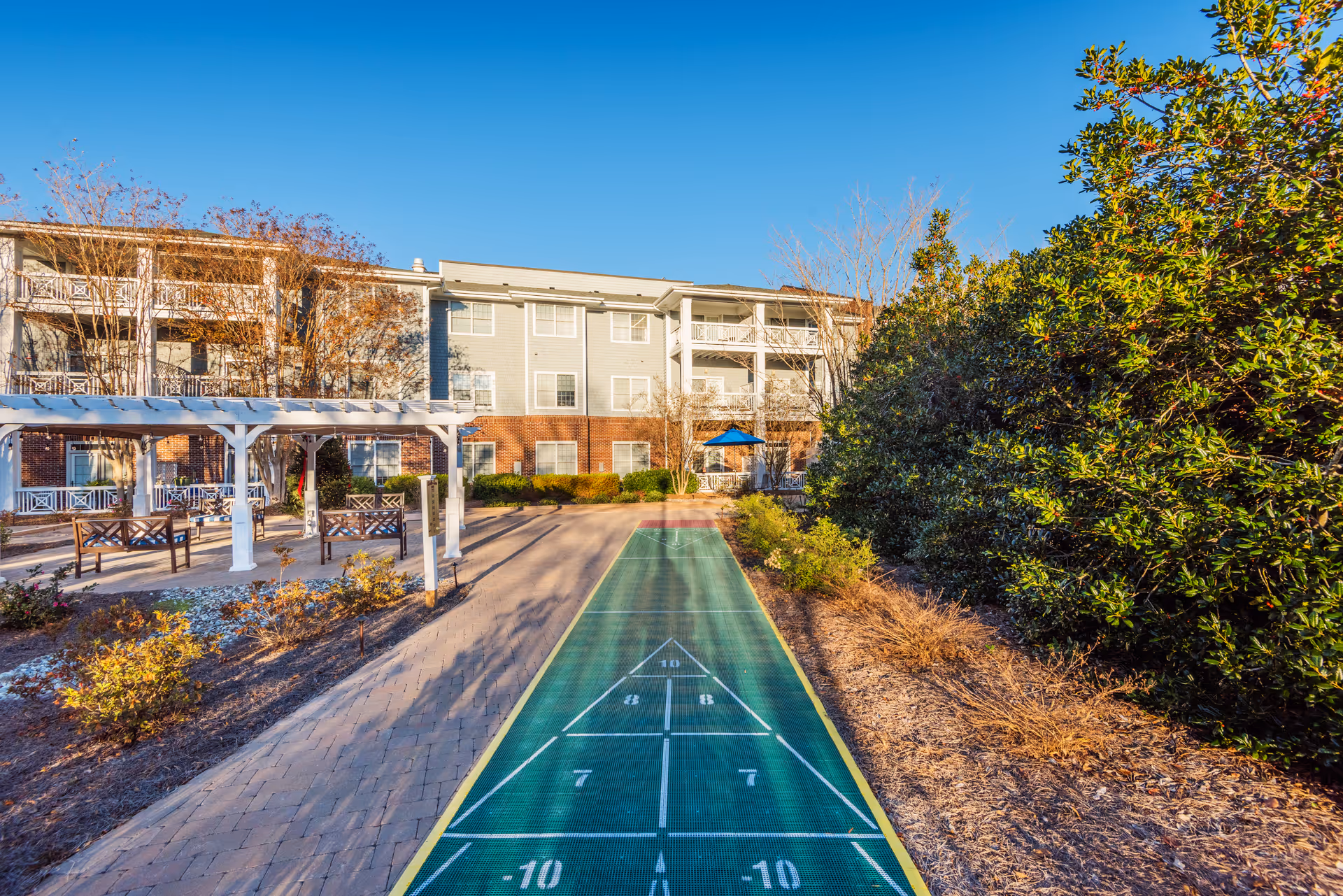 Outdoor shuffleboard court with a pergola and benches on the left side, surrounded by landscaped bushes and trees, with a multi-story residential building in the background under a clear blue sky.