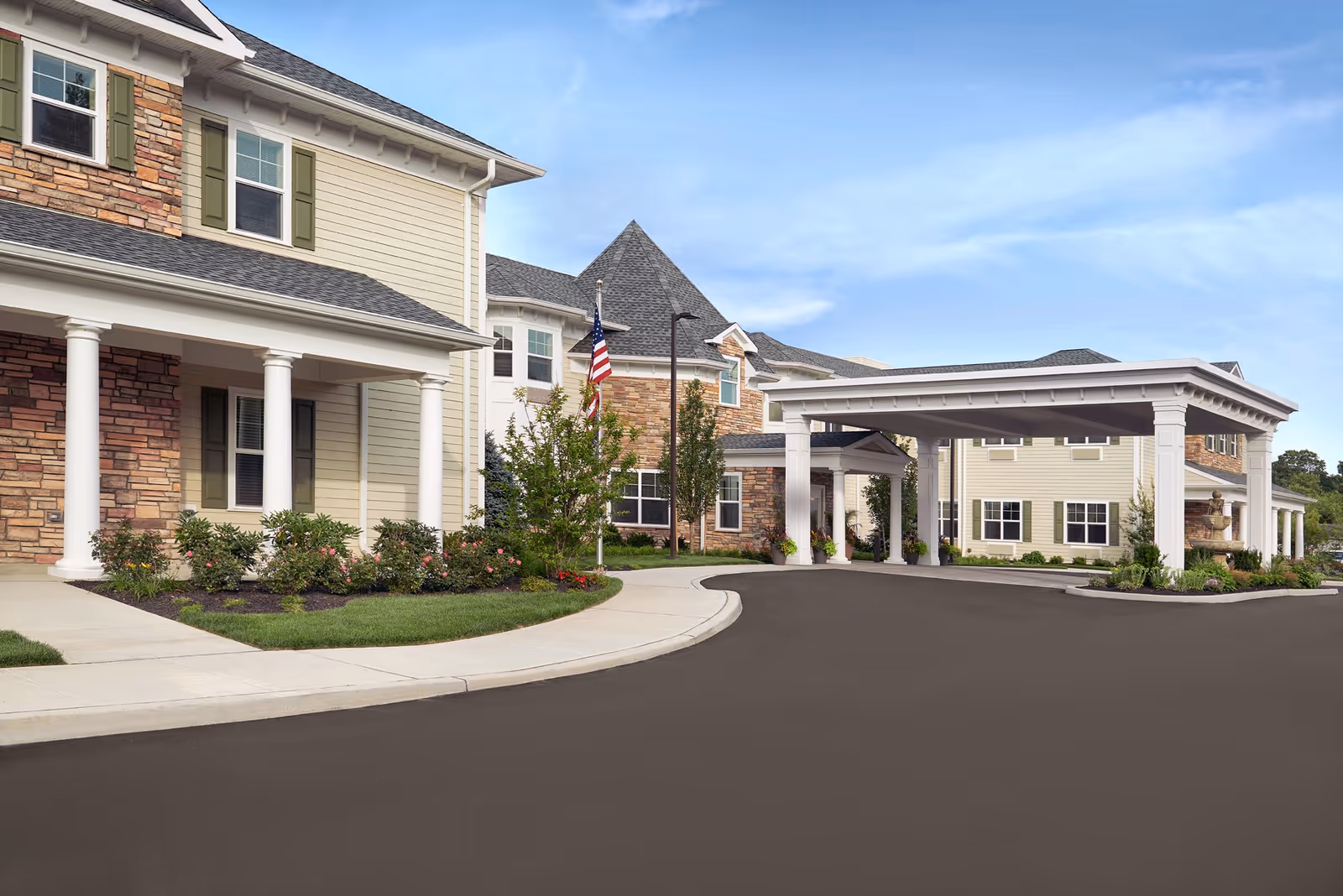 Exterior view of The Bristal Assisted Living at Sayville showing a large covered entrance with white columns, beige siding, stone accents, landscaped bushes, and an American flag near the building under a clear blue sky.