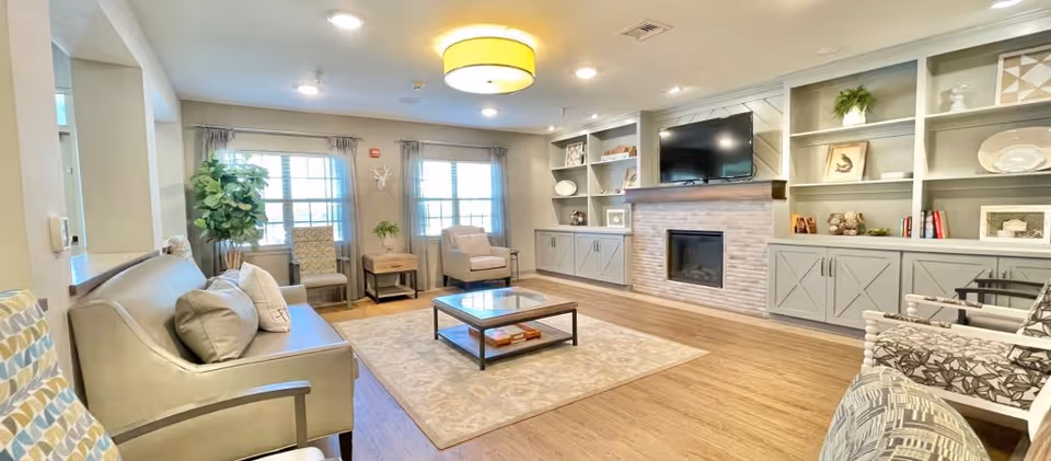 A bright and spacious living room with wooden flooring, a beige sofa with cushions, patterned armchairs, a glass coffee table on a light-colored rug, built-in shelves with decorative items, a wall-mounted flat-screen TV above a modern fireplace, and large windows with curtains allowing natural light to fill the room.